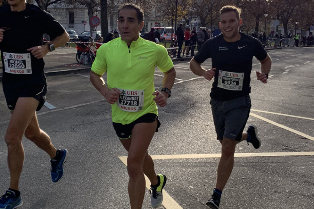Trois coureurs participent à une course sur route, portant des dossards numérotés. L'un d'eux, en t-shirt jaune fluorescent, court aux côtés de deux participants en noir, sous un ciel ensoleillé.