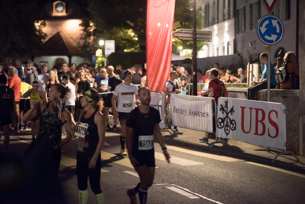 Participants à une course à pied urbaine en soirée (Demi de Jussy), passant devant des barrières affichant les logos de Berney Associés et UBS.