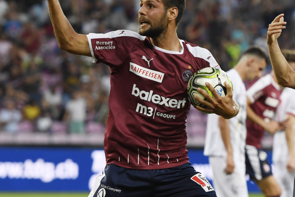 Joueur de football en maillot grenat (servette FC) tenant un ballon lors d'un match, avec d'autres joueurs visibles à l'arrière-plan sur le terrain