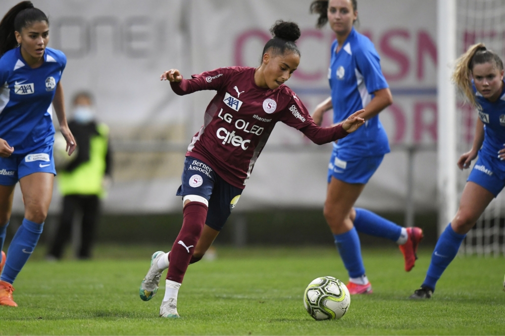 Joueuse de football en maillot grenat (Servette FC Chênois féminin) dribblant le ballon sur un terrain , entourée d'adversaires en tenue bleue lors d'un match.