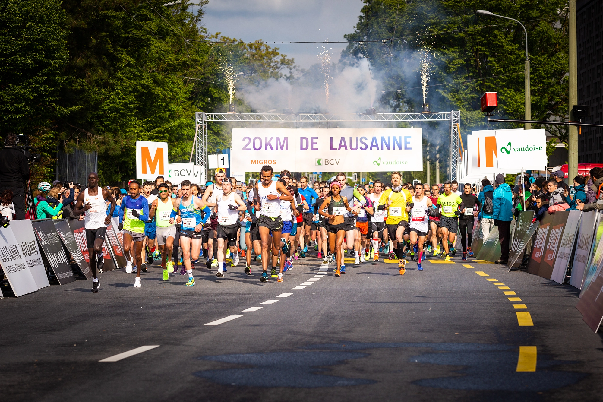 Départ d'une course des 20 km de Lausanne, avec un grand groupe de coureurs s'élançant sur la route sous l'arche de départ.