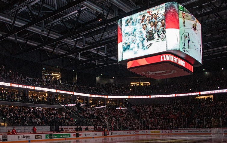 Patinoire de la vaudoise Arena remplie de spectateurs pendant un match de hockey du LHC, avec un grand écran suspendu affichant des joueurs et le message "l'union fait la force".