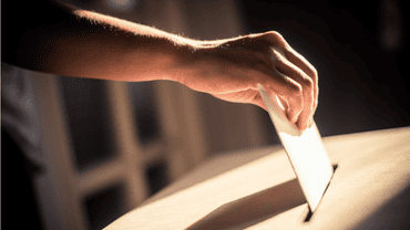 Main glissant une enveloppe dans une urne en cartons, illustrant le geste de vote ou de dépôt de bulletin.